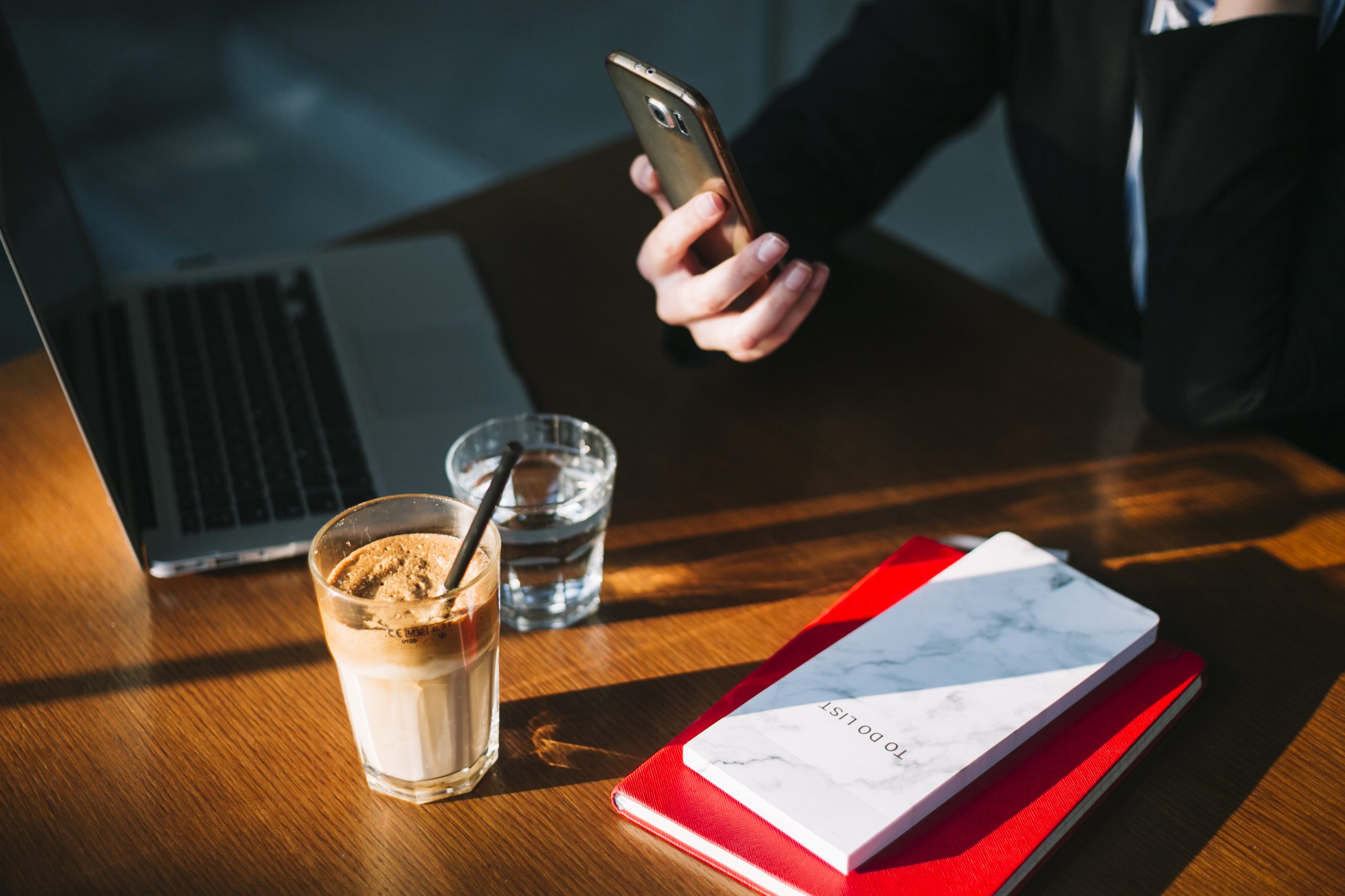 businesswoman-s-hand-using-cellphone-with-laptop-chocolate-milkshake-books-wooden-desk